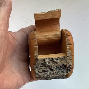 May include: A small, hand-carved wooden box with a hinged lid. The exterior features a natural, rough bark texture on the front, while the sides and interior are smooth, light-toned wood. The box has a rectangular opening and a vertical lid.