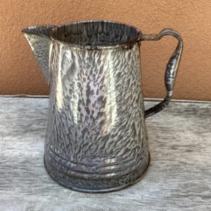 May include: A metal pitcher featuring a gray and white mottled enamel finish. The vessel has a wide circular opening, a prominent pouring spout on one side, and a curved handle attached to the opposite side. The base has three horizontal ridges.