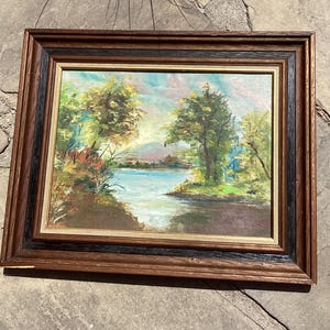 May include: A landscape oil painting in a dark brown and black wooden frame. The artwork depicts a calm blue lake surrounded by green trees and foliage under a soft sky, with distant hills visible in the center of the composition.