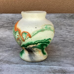 May include: A small, bulbous ceramic vase with a matte, off-white base featuring swirling, marbled patterns in shades of forest green, bright orange, and charcoal black. The vessel has a narrow neck, a flared rim, and a rounded body that tapers down.