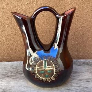 May include: A glossy, dark brown ceramic vase with a handle and a decorative sun design. The vase has a unique shape with two spouts and a central handle. The sun design features a cross and turquoise accents.