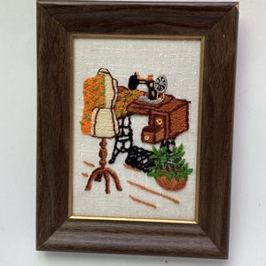 May include: A framed embroidery piece on off-white fabric featuring a brown wooden sewing table with a black sewing machine, a dress form draped with orange and green floral fabric, and a green potted plant, all enclosed in a dark wood frame.