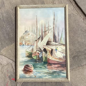 May include: Watercolor painting of a harbor scene with sailboats and a distant mosque. The boats are painted in shades of green, red, and white, with tall masts. The painting is framed in a gold-colored frame.