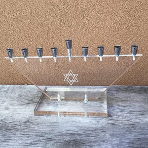 May include: Clear acrylic menorah with nine candle holders and a Star of David emblem. The base is rectangular, and the candle holders are silver-colored. The menorah is designed for the Jewish holiday of Hanukkah.