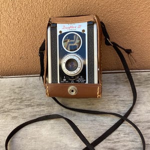 May include: A vintage Kodak Duaflex III camera with a brown leather case and a black strap. The camera has a black and silver face with the Kodak logo and lens details. The strap is looped in a figure-eight shape.