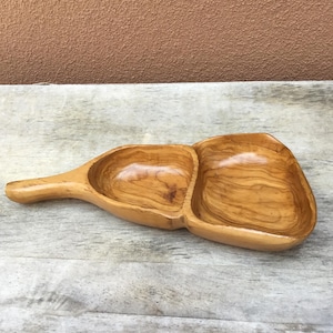 May include: A wooden serving tray with two compartments and a handle. The tray is made of light-colored wood with a natural grain pattern. The compartments are oval-shaped and appear to be smooth and polished. The handle is long and slender.