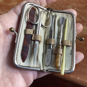 May include: A vintage manicure set in a small, silver-framed case. The set includes small scissors, tweezers, a cuticle pusher, and a yellow nail stick. The tools are secured with leather straps against a beige fabric background.