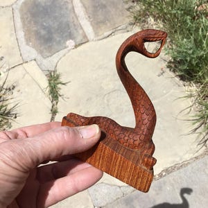 Puede incluir: Escultura de madera tallada de una serpiente con la boca abierta. La serpiente es de color marrón rojizo y tiene escamas detalladas. La escultura se sostiene en una mano sobre un fondo de piedra y hierba.