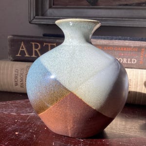 May include: A handcrafted ceramic vase with a rounded body and a narrow neck. The vase features a color-blocked design with sections of blue, white, brown, and rust. The vase is displayed on a wooden surface, with books in the background.