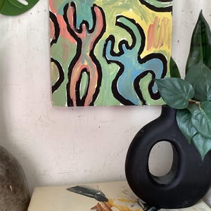 Puede incluir: Pintura abstracta cuadrada con un dise&ntilde;o colorido. La obra presenta figuras en rosa, azul y verde, delineadas en negro, sobre un fondo amarillo y verde. La pintura est&aacute; montada en una pared blanca.