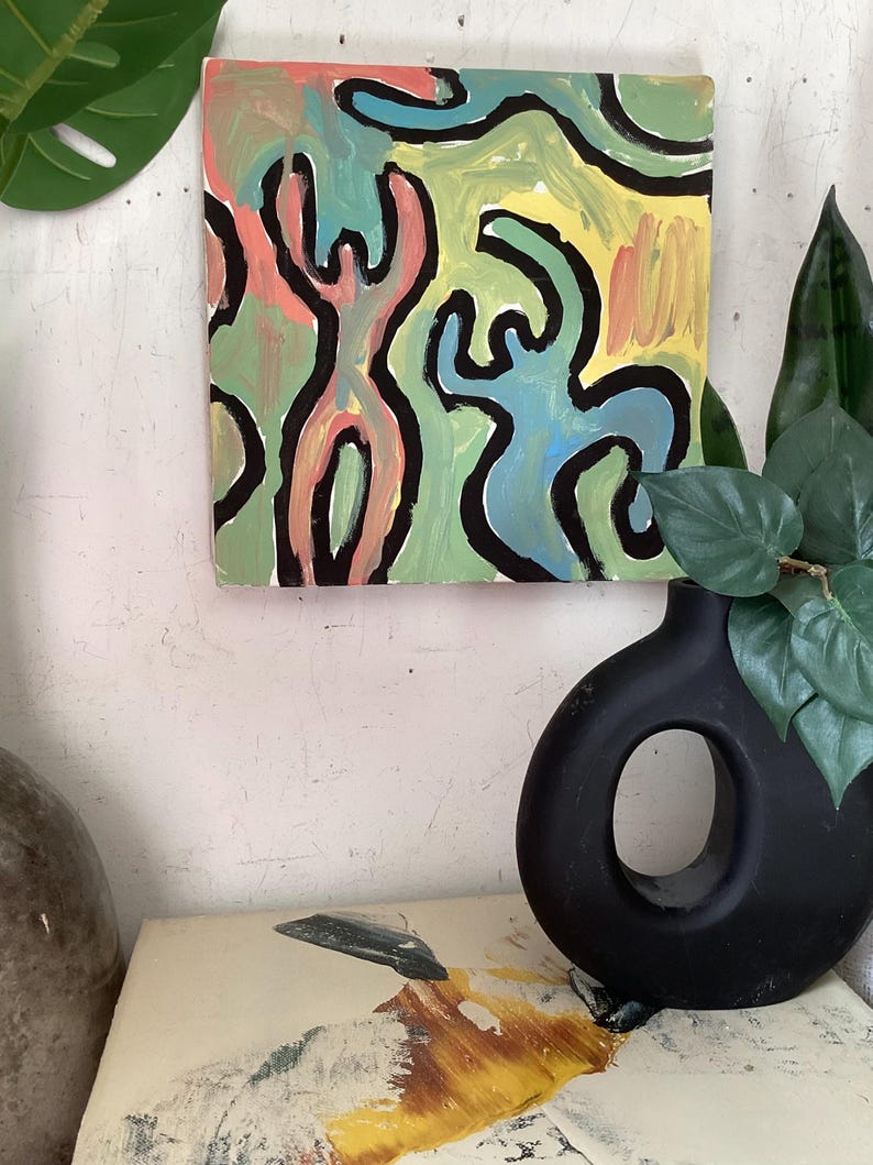 Puede incluir: Pintura abstracta cuadrada con un dise&ntilde;o colorido. La obra presenta figuras en rosa, azul y verde, delineadas en negro, sobre un fondo amarillo y verde. La pintura est&aacute; montada en una pared blanca.