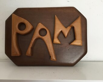 May include: Wooden sign with the name "PAM" carved in a light brown color, set against a dark brown, octagonal background. The letters are stylized and have a mid-century modern aesthetic. The sign is displayed on a white shelf.