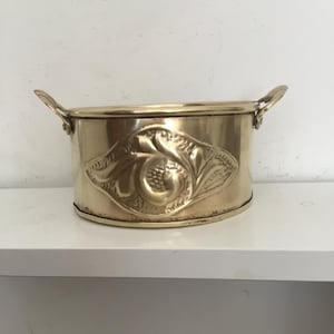 May include: A small, oval-shaped brass planter with two handles. The planter has an embossed floral design.