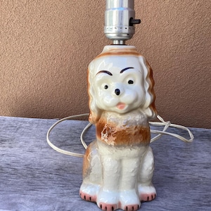 May include: A ceramic dog-shaped table lamp with a white, brown, and pink color scheme. The lamp features a seated dog design with a light socket at the top. The lamp has a white cord.