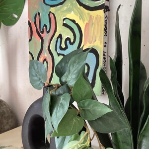 Puede incluir: Una pintura abstracta con un dise&ntilde;o colorido de formas amarillas, verdes, azules y rojas, delineadas en negro. La obra de arte est&aacute; montada en una pared, con un jarr&oacute;n negro y plantas verdes en primer plano. El texto "ART CONCEPT by KREBS" es visible.