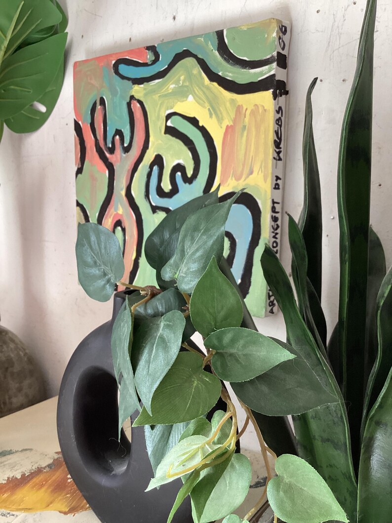 Puede incluir: Una pintura abstracta con un dise&ntilde;o colorido de formas amarillas, verdes, azules y rojas, delineadas en negro. La obra de arte est&aacute; montada en una pared, con un jarr&oacute;n negro y plantas verdes en primer plano. El texto "ART CONCEPT by KREBS" es visible.