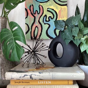 Puede incluir: Arreglo decorativo con un jarr&oacute;n negro, una escultura met&aacute;lica en forma de estrella y una pila de libros. El fondo presenta una colorida pintura abstracta y follaje verde. La palabra "LISTEN" es visible.