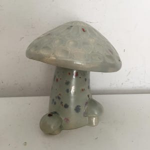 May include: A ceramic mushroom figurine with a light green glaze and a speckled pattern of red, blue, and white. The mushroom has a rounded cap and a thick stem with a bulbous base.