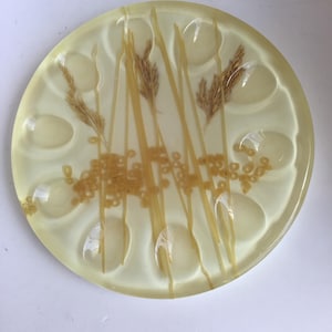 May include: A round, yellow, resin egg holder with six indentations. The holder has dried grass and spaghetti noodles embedded in the resin.
