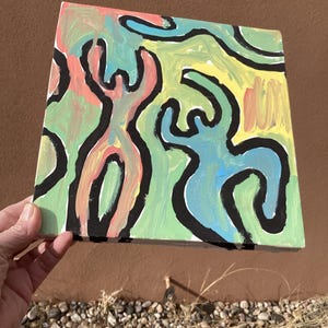 Puede incluir: Arte en lienzo cuadrado con figuras abstractas delineadas en negro. Las figuras est&aacute;n rellenas de rosa, azul y verde, sobre un fondo amarillo y verde. La obra de arte est&aacute; sostenida por una mano. La pintura es cuadrada.