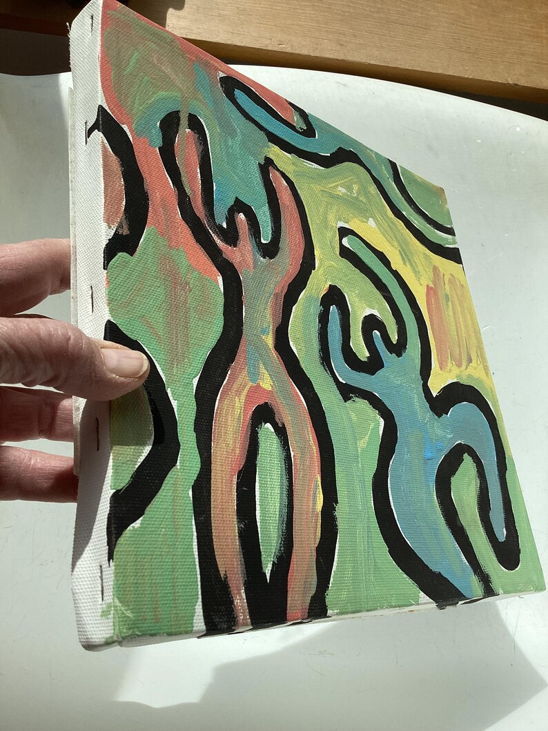 Puede incluir: Lienzo de arte abstracto con formas coloridas delineadas en negro. La obra de arte presenta figuras en tonos verdes, amarillos, azules y coral. El lienzo est&aacute; estirado y grapado a un marco de madera, listo para exhibir. La pintura est&aacute; sostenida por una mano.