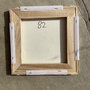 Puede incluir: Parte trasera de una obra de arte en lienzo cuadrado con marco de madera. El lienzo est&aacute; estirado y grapado al marco. El n&uacute;mero "82" est&aacute; escrito a mano en el lienzo. Tiras de tela blanca est&aacute;n adheridas al marco.