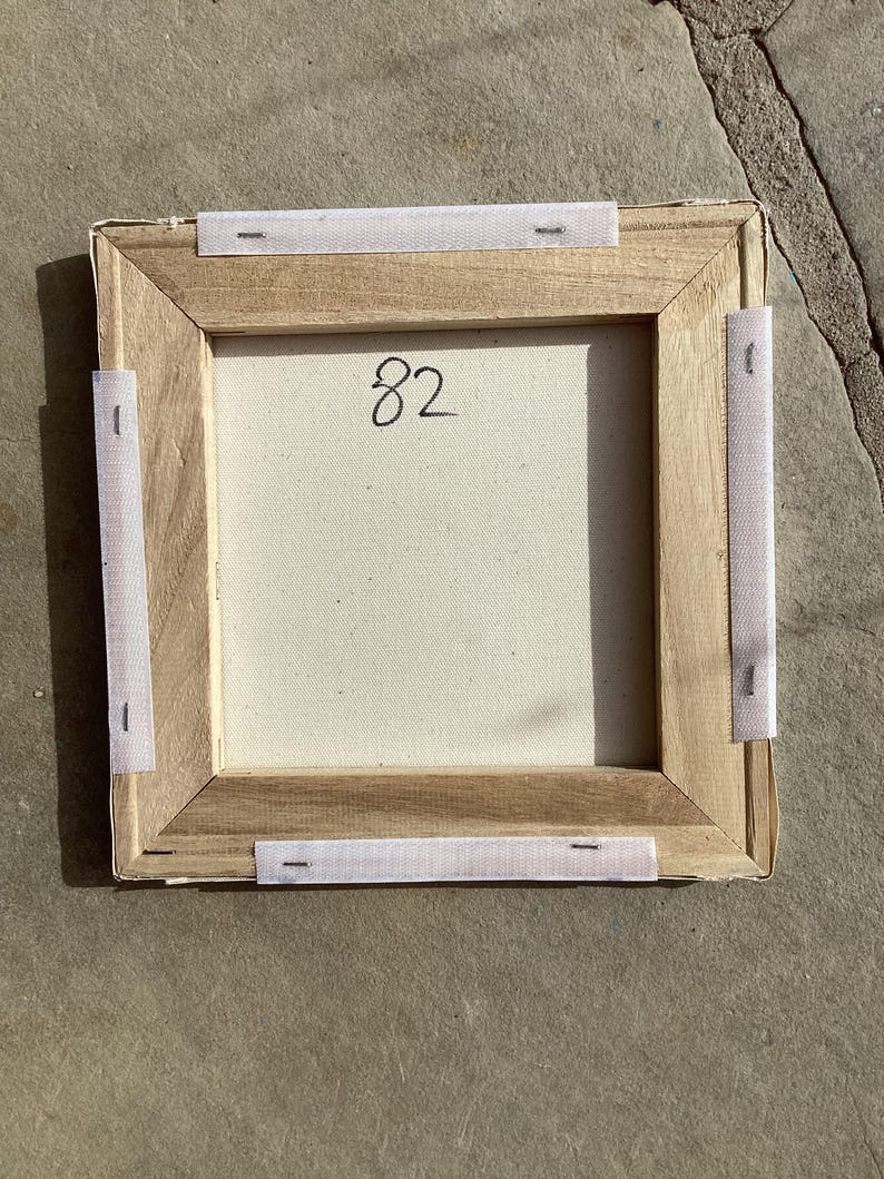 Puede incluir: Parte trasera de una obra de arte en lienzo cuadrado con marco de madera. El lienzo est&aacute; estirado y grapado al marco. El n&uacute;mero "82" est&aacute; escrito a mano en el lienzo. Tiras de tela blanca est&aacute;n adheridas al marco.