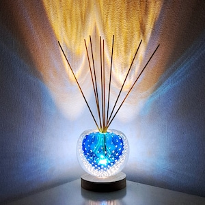 Könnte beinhalten: Ein dekorativer Glasdiffusor mit blauem Innenraum und weißen Akzenten, der auf einem Holzsockel steht. Braune Schilfrohre erstrecken sich aus der Glaskugel. Der Hintergrund ist eine strukturierte Wand mit goldenen Lichtmustern.