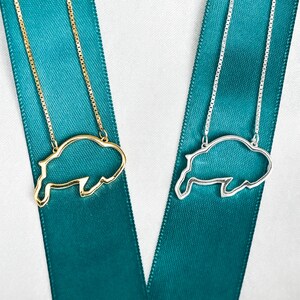 May include: Two necklaces with buffalo silhouette pendants. One necklace has a gold pendant and the other has a silver pendant. Both necklaces are on a teal ribbon.