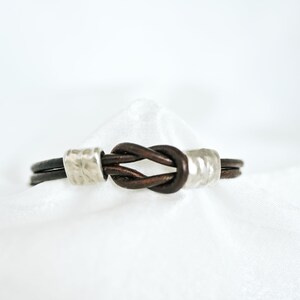 May include: A brown leather bracelet with a knot design. The bracelet features a knot in the center and is accented with silver-colored metal cylinders. The bracelet is displayed on a white background.