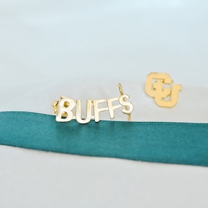 May include: Gold-colored metal earrings with the word "BUFFS" and the letters "CU" on a white background with a green ribbon.