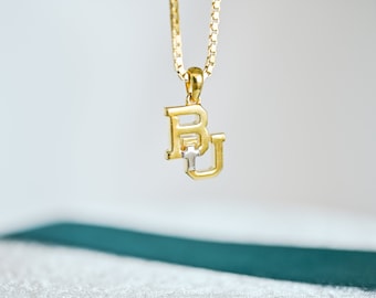 May include: A gold chain necklace with a gold pendant featuring the letters "BU" in a bold font. The pendant has a small cross in the center.