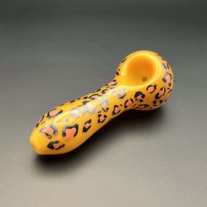 Puede incluir: Una pipa de vidrio amarilla con un diseño de estampado de leopardo rosa y negro.