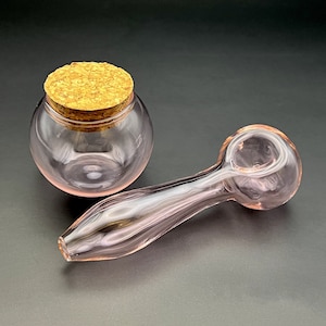 Op de afbeelding: Een roze glazen pijp met een ronde kom en een lange, gebogen steel. De pijp ligt naast een roze glazen pot met een kurken deksel.