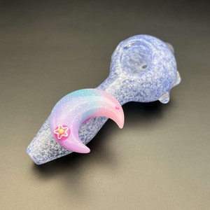 May include: A blue glass pipe with a crescent moon design. The moon is made of iridescent glass with a pink and blue color gradient. A small flower with a gold center is attached to the moon.