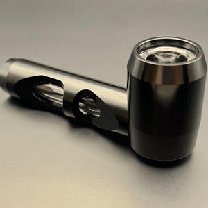 May include: A metallic pipe with a cylindrical bowl and a curved stem. The bowl is black, while the stem is silver with a cut-out design. The pipe is designed for smoking and rests on a gray surface.