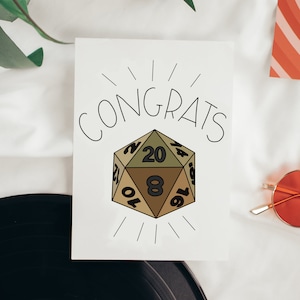 May include: A white card with the word "CONGRATS" written in black. A black and white illustration of a 20-sided die with the numbers 2, 4, 6, 8, 10, 12, 14, 16, 18, 20, 1, 3, 5, 7, 9, 11, 13, 15, 17, and 19. The die is green, brown, and tan.