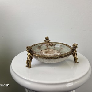 May include: An antique decorative bowl with a gold-colored rim and cherub figures on each side. The bowl has a cream-colored base with a brown and green floral design. The bowl is sitting on a white pedestal.
