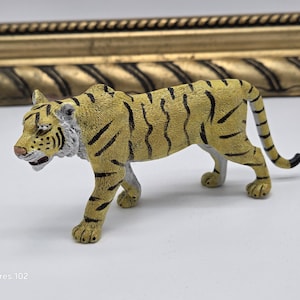 Puede incluir: Figura de tigre amarillo y negro con un diseño detallado. El tigre está en posición de caminar, con la cola curvada. La figura tiene un aspecto texturizado y está sobre un fondo blanco. El texto "Sculptures 102" es visible.