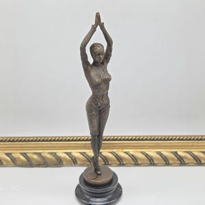 May include: A bronze-coloured sculpture of a woman with arms raised above her head. The figure is detailed with etched patterns and stands on a round, dark base. The sculpture is set against a gold-framed background.