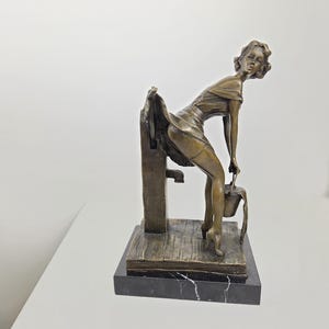 Op de afbeelding: Bronzen beeldhouwwerk van een vrouw die bukt en een emmer vasthoudt. De figuur bevindt zich in de buurt van een waterpomp. Het beeldhouwwerk is gemonteerd op een zwarte marmeren voet. De vrouw draagt een jurk.