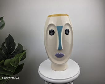 May include: A cream-colored ceramic vase shaped like a stylized human face. It features a blue vertical nose bridge, two black circular eyes, and purple lips. The top rim is accented with a mustard yellow band. The text Sculptures.102 is visible.
