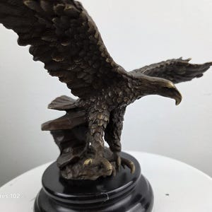 May include: Bronze eagle sculpture with outstretched wings, mounted on a black base. The eagle's feathers are detailed, and the sculpture has a weathered appearance. A decorative item.
