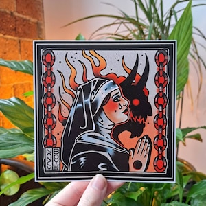 May include: A black and white illustration of a nun with a tear on her cheek, praying with her hands clasped together. Behind her is a red devil with horns, surrounded by flames. The image is framed by a black border and a chain with red links.