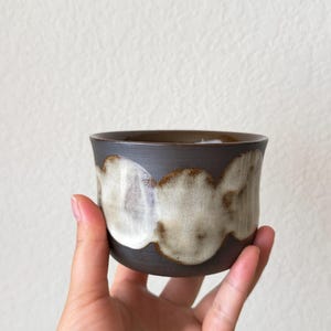 May include: A handmade ceramic cup with a dark brown exterior and a cream-colored, cloud-like design around the middle. The cup is held in a person's hand, showcasing its unique shape and texture. The cup is likely used for drinking.