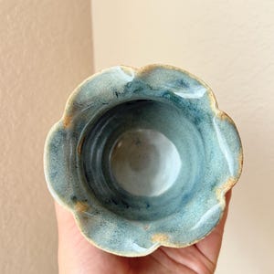 May include: A handmade ceramic bowl with a scalloped rim. The bowl is glazed in shades of blue and green, with a light brown rim. The interior is a lighter blue. The bowl is held in a hand.