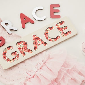 May include: A wooden name puzzle with the word "GRACE" cut out. The letters are red and white and are scattered around the puzzle. The puzzle is on a pink background with a pink dress and pink shoes.