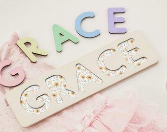 May include: A wooden name puzzle with the name "GRACE" spelled out in cutout letters. The letters are filled with a floral pattern featuring yellow sunflowers and green leaves. The puzzle is on a white surface with a pink baby outfit and pink baby shoes.
