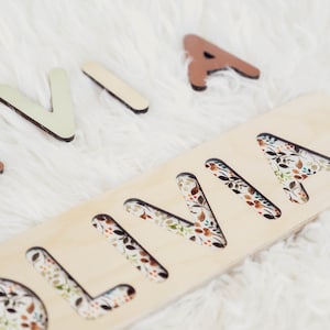 May include: Wooden puzzle with the name "OLIVIA" cut out. The letters are surrounded by a floral pattern. The letters are separate and can be placed in the cutouts.