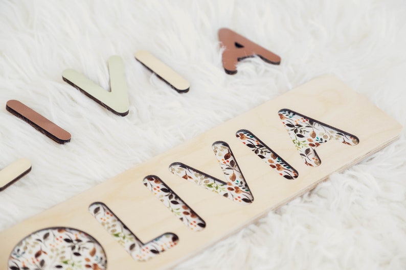 May include: Wooden puzzle with the name "OLIVIA" cut out. The letters are surrounded by a floral pattern. The letters are separate and can be placed in the cutouts.